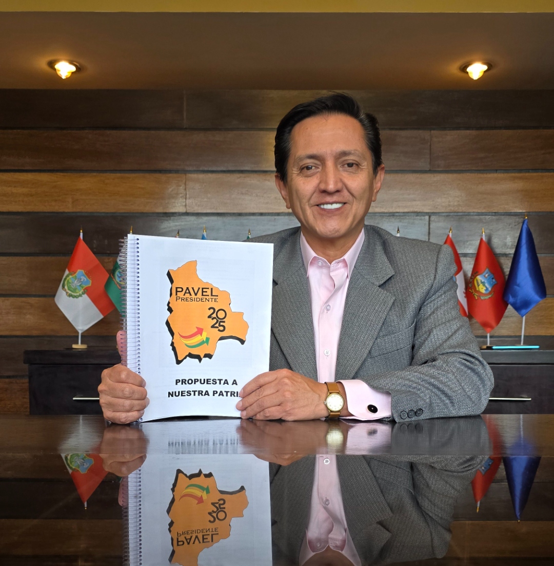 Pavel Aracena sonriendo en una oficina con banderas departamentales de Bolivia, sosteniendo el documento de propuesta Pavel Presidente 2025